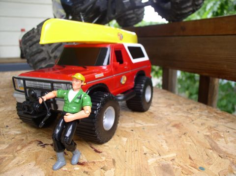 1979 Ford Bronco - A toy Ford Bronco is outfitted for adventure with a canoe and a figure, while another toy truck looms overhead.