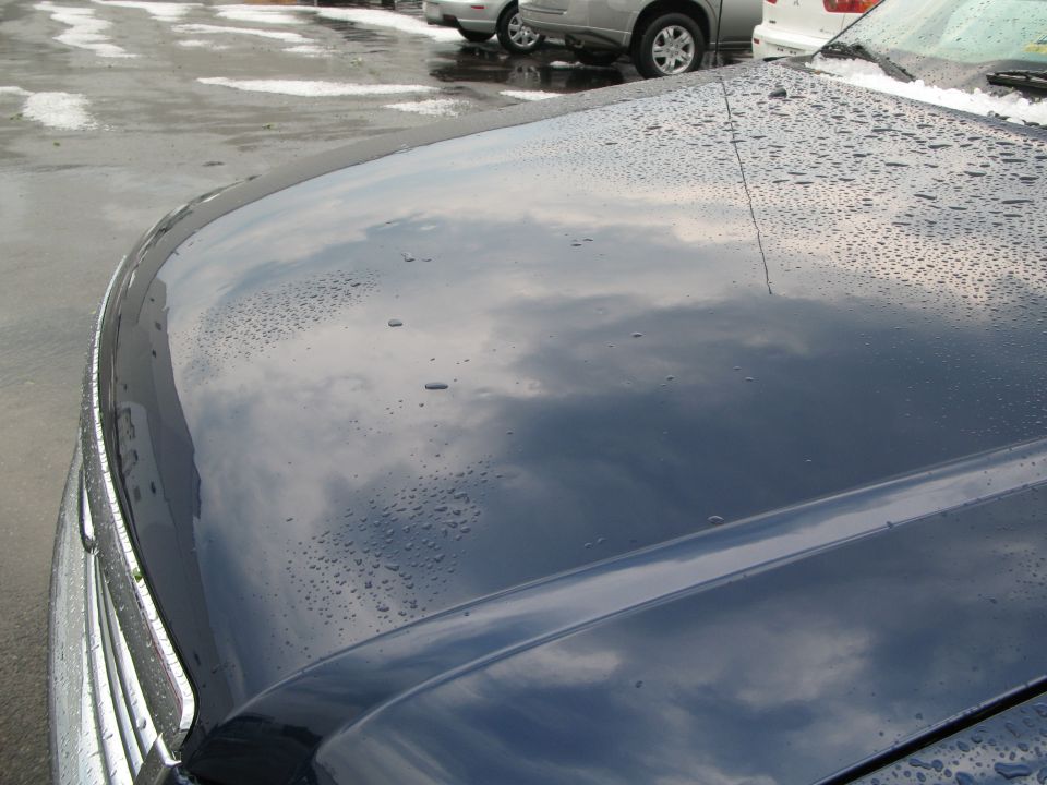 Dark blue car hood with water droplets and reflection of clouds. Other vehicles visible in background