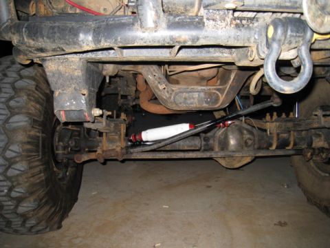 1996 Ford Bronco - Underside view looking up at a lifted 4x4's front axle and steering, showing the steering box, pitman arm, drag link and a white Rancho-sty…