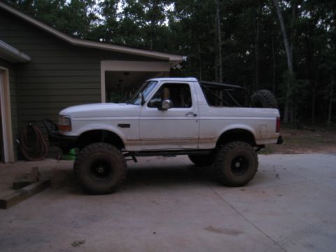 1996 Ford Bronco - Side profile of a lifted white Ford Bronco with the fiberglass rear top removed, exposing a black roll bar, parked on a concrete driveway b…