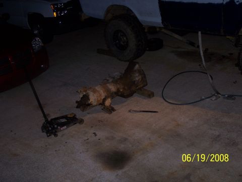 1979 Ford F-150 - A transmission part lies on the ground next to a jack, with vehicles in the background.