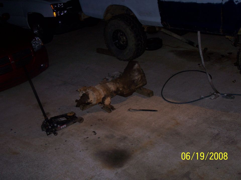 Transmission part on concrete floor with a jack and part of a red car and a truck in the background