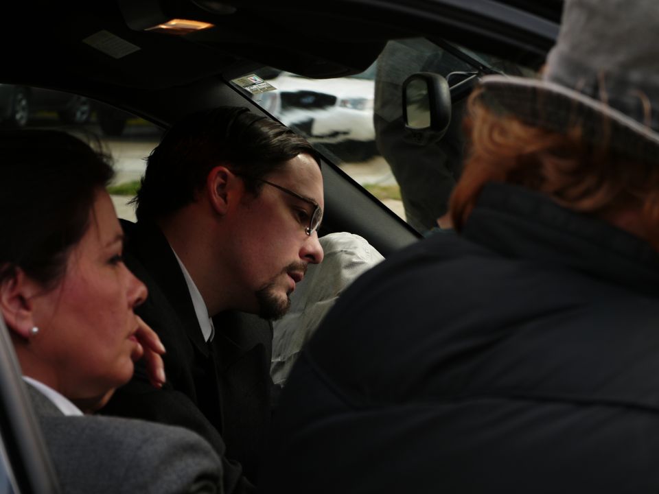 People inside a car, one man in glasses looking forward, another woman to his left, and a third person's back visible