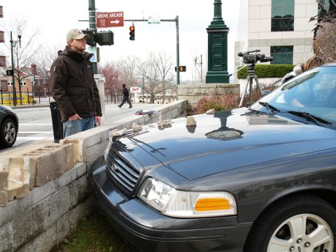1985 Ford LTD Crown Victoria - A dark sedan is parked at an angle next to a concrete barrier, with a man standing nearby.