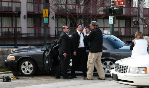 1985 Ford LTD Crown Victoria - A dark sedan, likely a 2004 Ford Crown Victoria, is seen with its door open as men in suits stand around it, with another vehicle nearby.