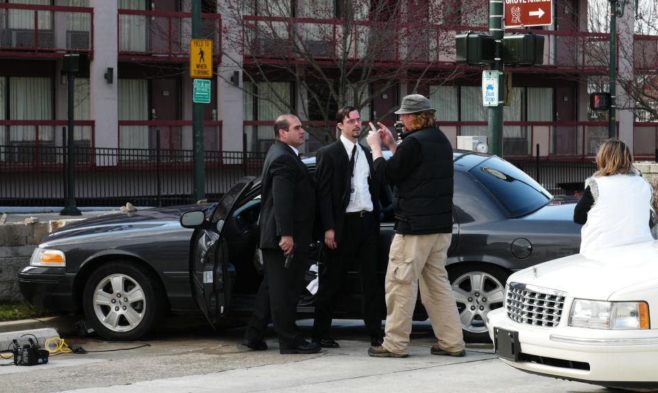 Dark sedan with open door, men in suits standing nearby. A white Cadillac is partially visible