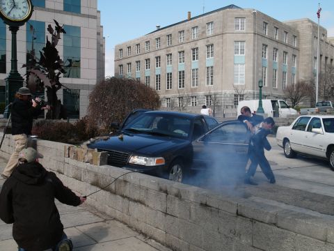 1985 Ford LTD Crown Victoria - A black Ford Crown Victoria is partially over a stone wall with smoke billowing out, as two men in suits interact with the vehicle.