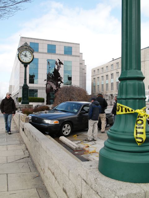 1985 Ford LTD Crown Victoria - A dark sedan is parked on a sidewalk next to a stone wall, with caution tape visible on a lamppost in the foreground.