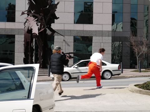 1985 Ford LTD Crown Victoria - A person with a camera appears to be filming another person running past a white car in front of a modern building.