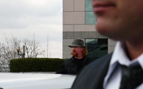1985 Ford LTD Crown Victoria - A man wearing a hat and vest stands outside a building, with a person in a suit visible in the foreground.