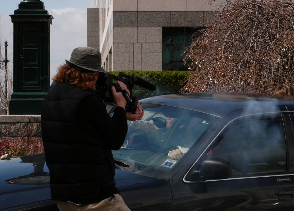 Black sedan with smoke billowing from the roof, viewed from the side with a cameraman filming
