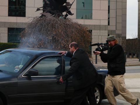 1985 Ford LTD Crown Victoria - A man in a suit opens the door of a dark sedan while a cameraman films the scene.