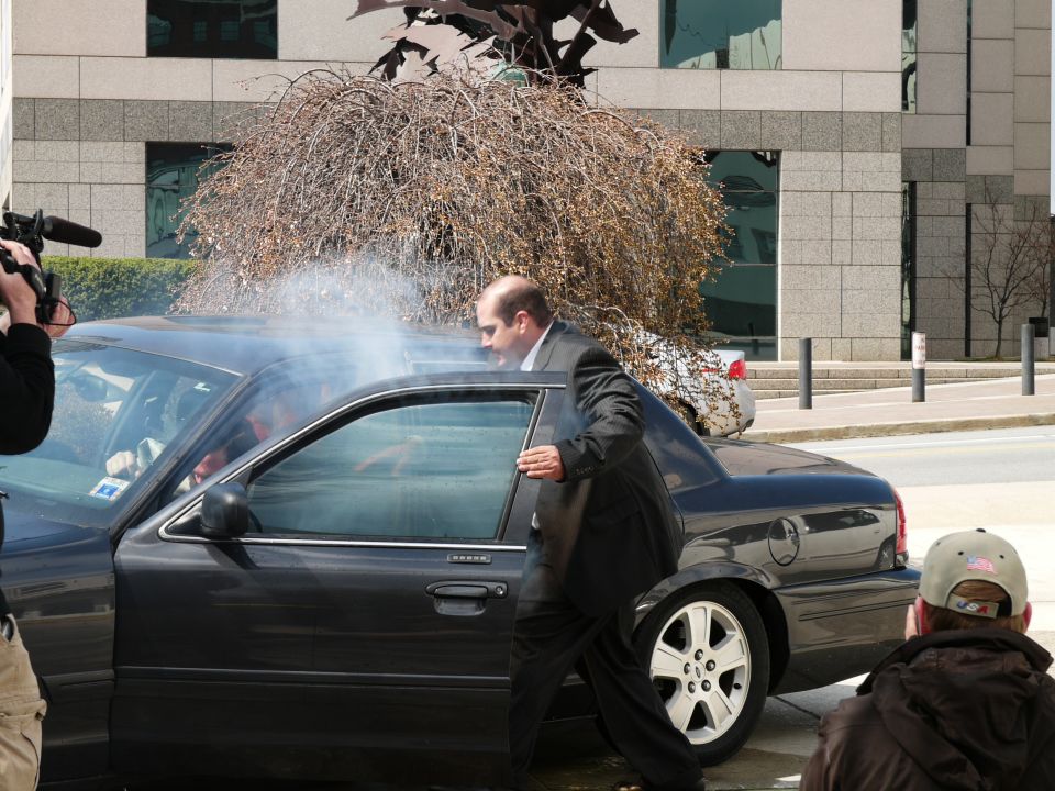 Man in suit exiting black sedan, smoke visible from door. Another car parked behind