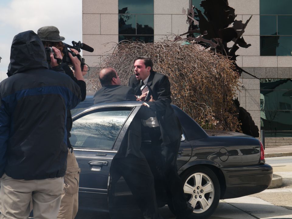 Filming a scene with two men in suits arguing next to a dark sedan, with a camera crew present