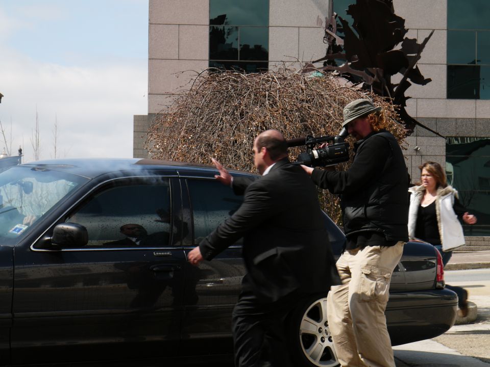 Black sedan side view with a man opening the door and a cameraman filming