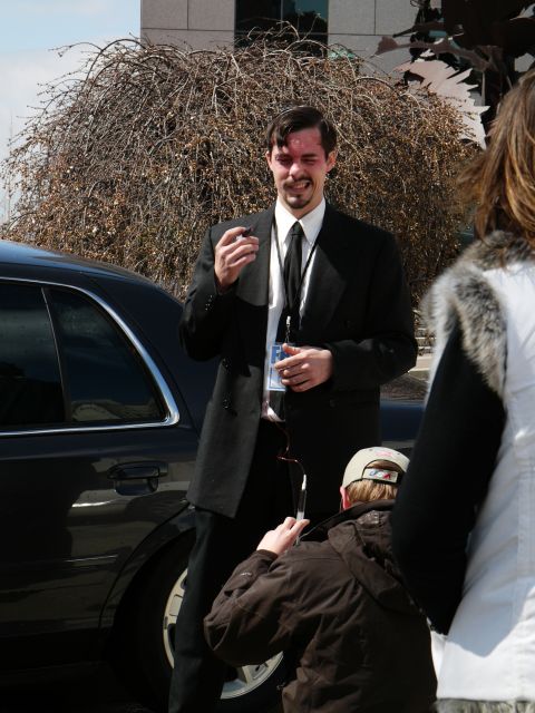 1985 Ford LTD Crown Victoria - A man in a suit holds a cigar while standing by a black sedan, as someone kneels to take a photo.