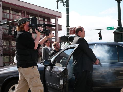 1985 Ford LTD Crown Victoria - A man with a gun stands by a dark sedan as smoke billows from the open door, with a cameraman filming.