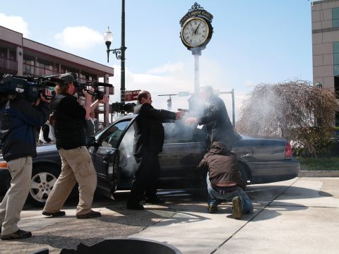 1985 Ford LTD Crown Victoria - A dramatic scene unfolds as two men point guns at each other beside a dark sedan, while news crews capture the action.