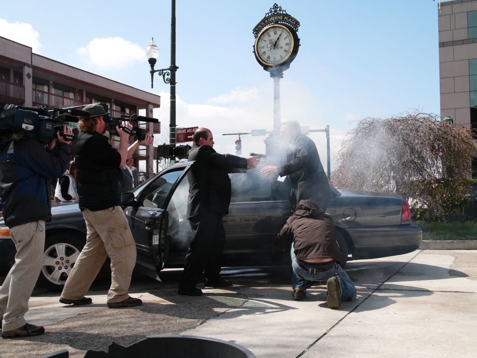 Dark sedan side view with open door, two men pointing guns, smoke, and news cameras filming