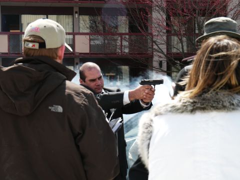1985 Ford LTD Crown Victoria - A man in a suit fires a handgun, creating a cloud of smoke. The scene appears to be outdoors.