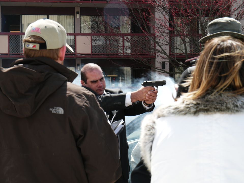 Man in suit fires handgun, smoke coming from barrel. People in foreground and background