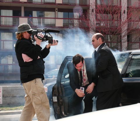 1985 Ford LTD Crown Victoria - A man in a suit exits a dark sedan amidst smoke, while a cameraman films the scene.