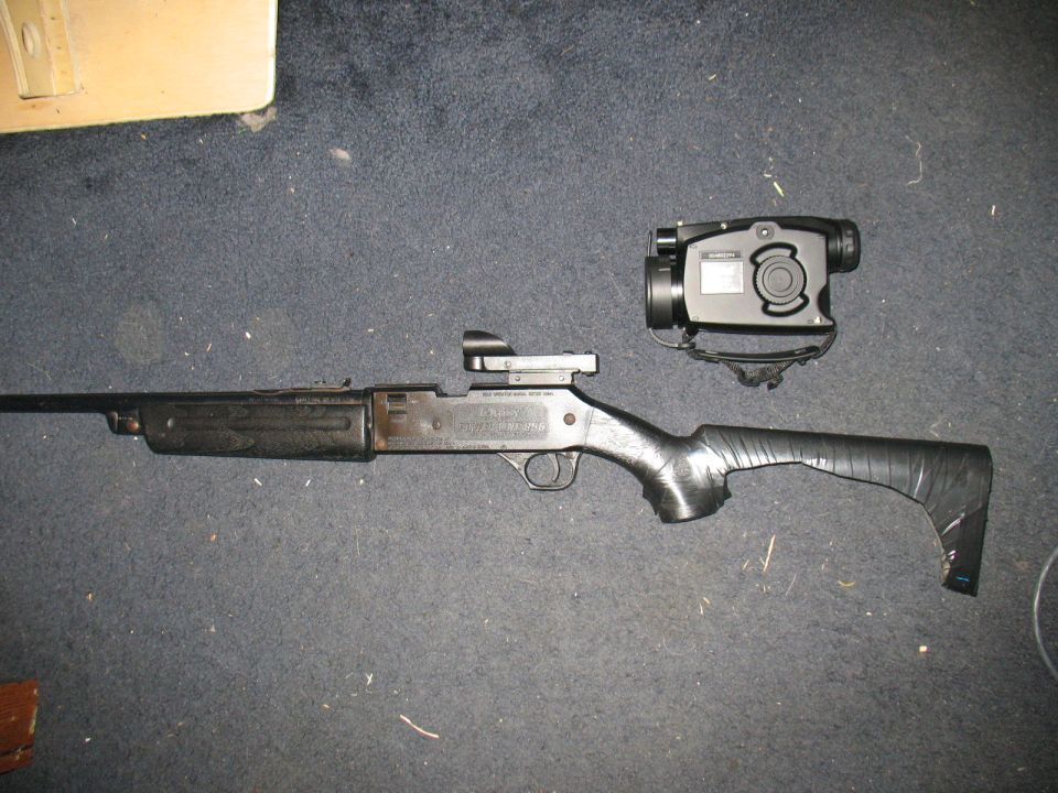 Black shotgun with a scope and a night vision device, lying on a dark carpet. The stock is wrapped in black tape