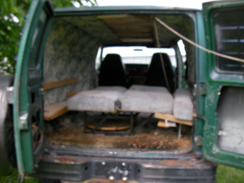 Green van interior with two front seats, two beds, and wooden shelves. The rear doors are open