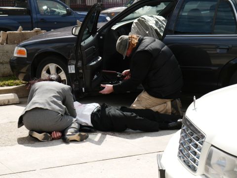 1985 Ford LTD Crown Victoria - A dark sedan is seen with its door open as people attend to a person lying on the ground, with another vehicle partially visible in the for…