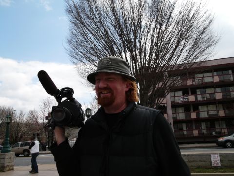 1985 Ford LTD Crown Victoria - A man holds a video camera and microphone in front of a building and bare trees.