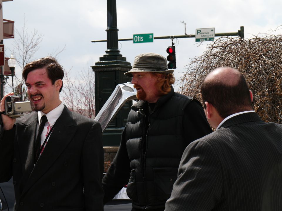Man with video camera films two other men on a street with a traffic light and street sign
