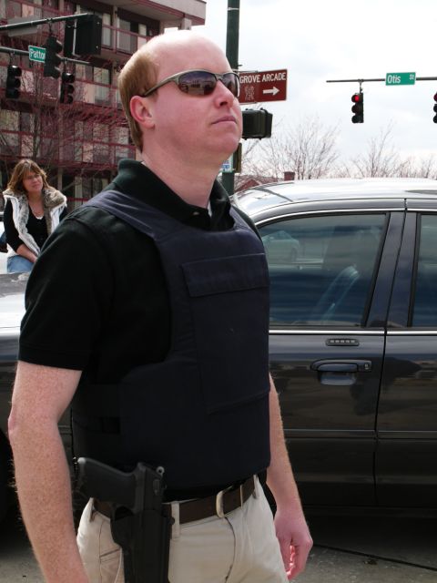 1985 Ford LTD Crown Victoria - A dark sedan is visible behind a man in a bulletproof vest.