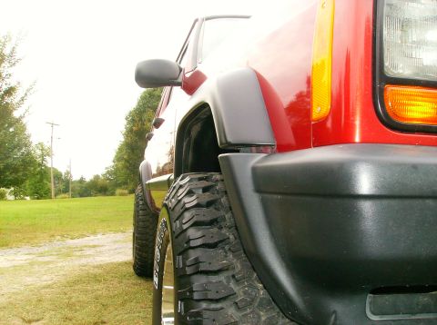 1998 Jeep Cherokee - Close-up of a red Jeep Cherokee's front fender and tire, showing off-road tires and black trim.