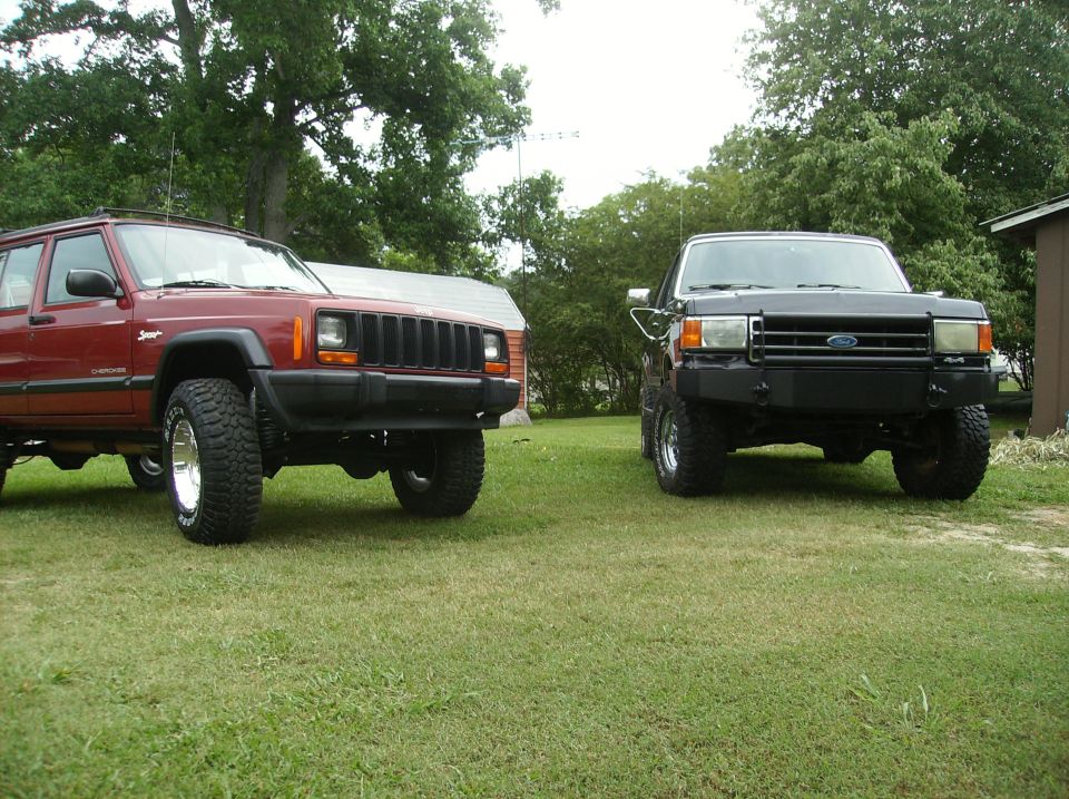 Red Jeep Cherokee and black Ford Bronco with off-road tires parked on grass.