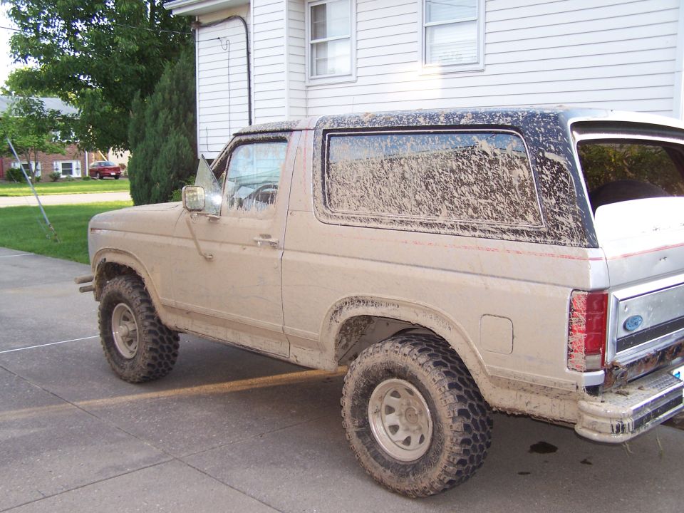 Mud-covered tan Ford Bronco SUV with off-road tires parked in a driveway, rear three-quarter view