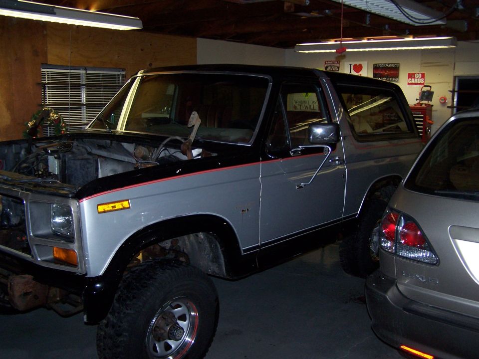 Silver and black Ford Bronco with open hood and off-road tires parked in a garage.