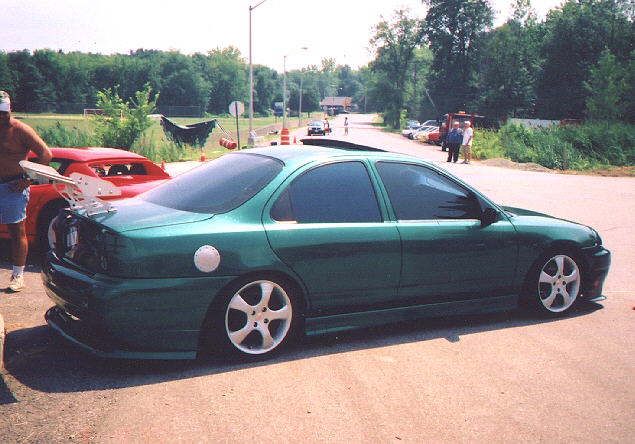 Green lowered sedan with white wheels and rear spoiler, side-rear view, parked outdoors