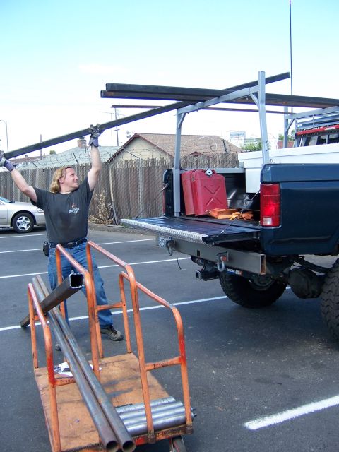 1991 Ford Bronco - Photograph of a person loading long steel tubing onto a pickup truck's rack in a parking lot.