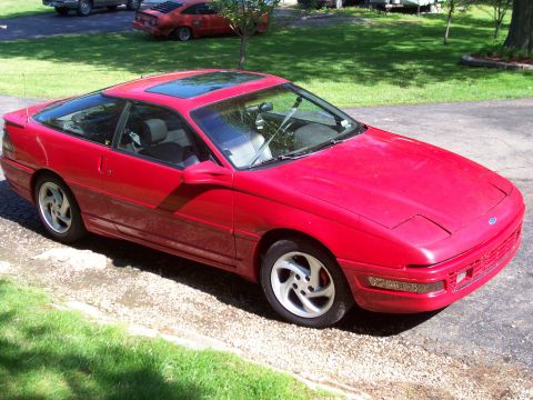 1989 Ford Probe - A red 1989 Ford Probe is parked on a gravel driveway next to a grassy lawn. The car has a sunroof and pop-up headlights.