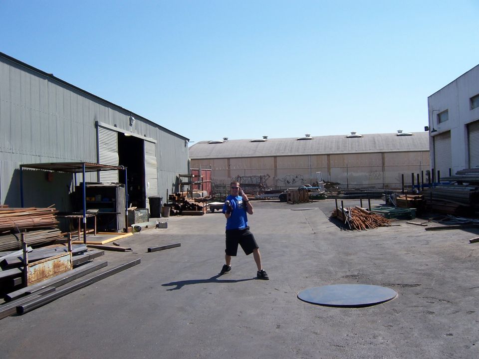 Person standing in a metal-supply yard with stacks of steel and warehouses