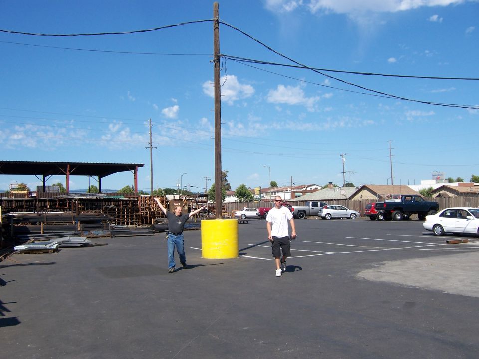 Two men in a paved metal-supply yard, one beside a yellow bollard