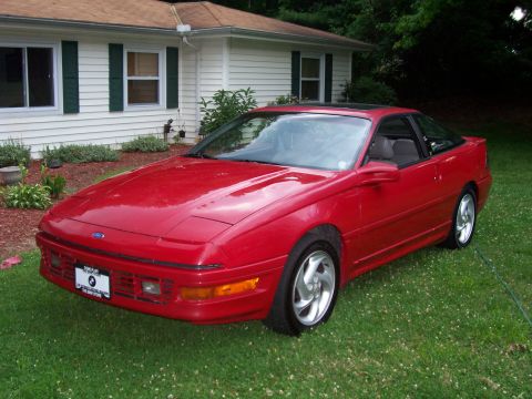 1989 Ford Probe - A red 1989 Ford Probe is parked on a grassy lawn in front of a white house with green shutters. The car is viewed from a 3/4 front angle, s…