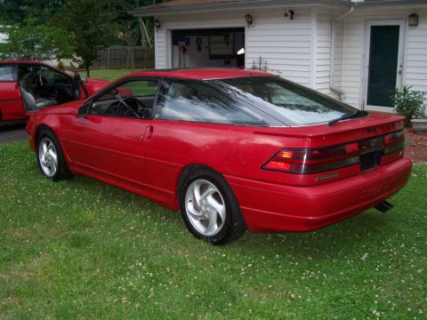 1989 Ford Probe - A red 1989 Ford Probe GT is parked on a grassy lawn in front of a white house with a garage. The car is viewed from the rear passenger side…