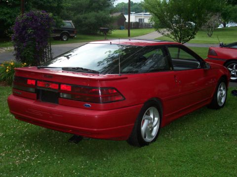 1989 Ford Probe - Rear three-quarter view of a red Ford Probe parked on a grassy lawn with a tree and bushes in the background. The car has a spoiler and a p…