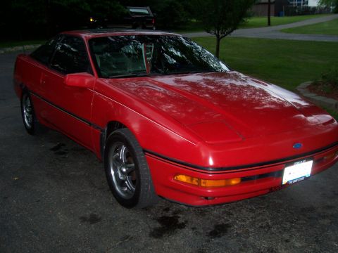 1989 Ford Probe - A red 1989 Ford Probe is parked on a wet asphalt driveway in front of a grassy lawn and trees. The car is viewed from a 3/4 front angle, sh…