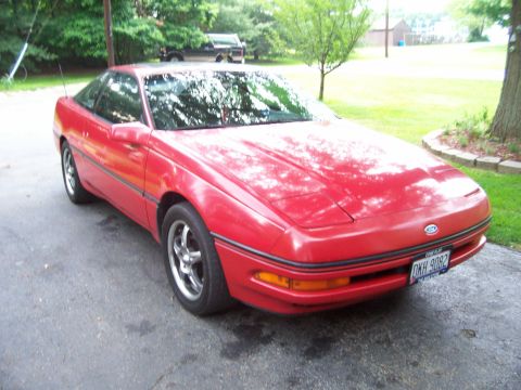 1989 Ford Probe - A red 1989 Ford Probe is parked on an asphalt driveway in front of a grassy area with trees. The car is viewed from a 3/4 front angle, show…