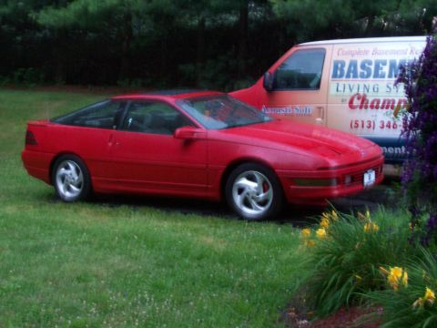 1989 Ford Probe - A red 1989 Ford Probe is parked on a grassy lawn next to a van. The car is viewed from the side with a 3/4 front angle, showing its sporty…