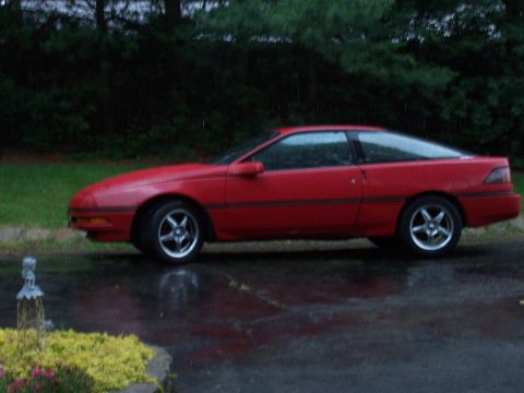 1989 Ford Probe - The side profile of a red 1989 Ford Probe is shown parked on a wet asphalt surface with a grassy area and trees in the background. The car…