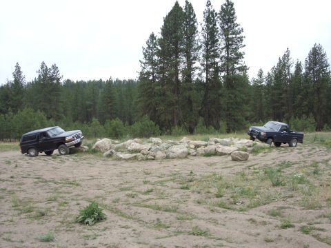 1988 Ford Bronco - Wide shot of a dirt clearing in a pine forest with two dark Ford Broncos parked at opposite ends of a long line of boulders, one with its h…