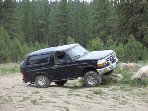 1988 Ford Bronco - Driver-side profile of a black full-size Ford Bronco with its hardtop on, front wheel perched up on a rock at the edge of a dirt clearing s…
